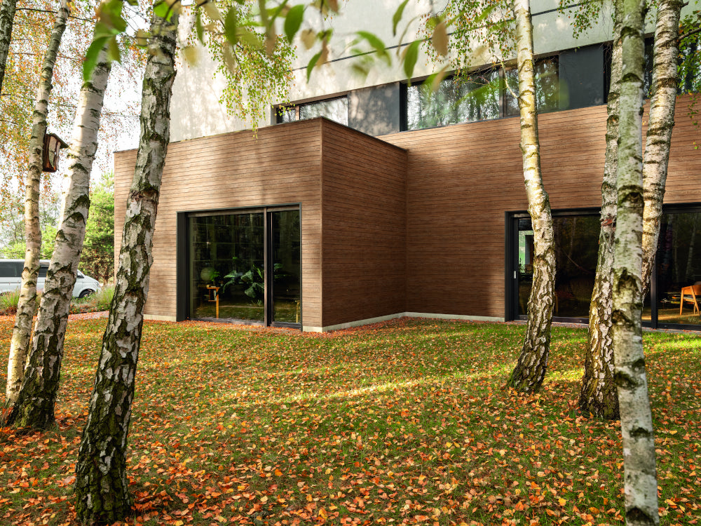 Modern house with wooden facade and large windows, surrounded by trees and grass with fallen leaves.