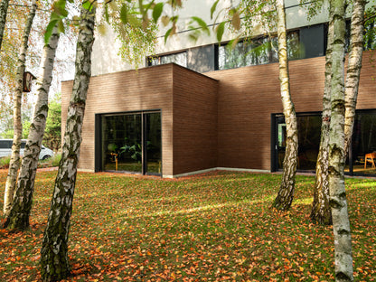 Modern house with wooden facade and large windows, surrounded by trees and grass with fallen leaves.